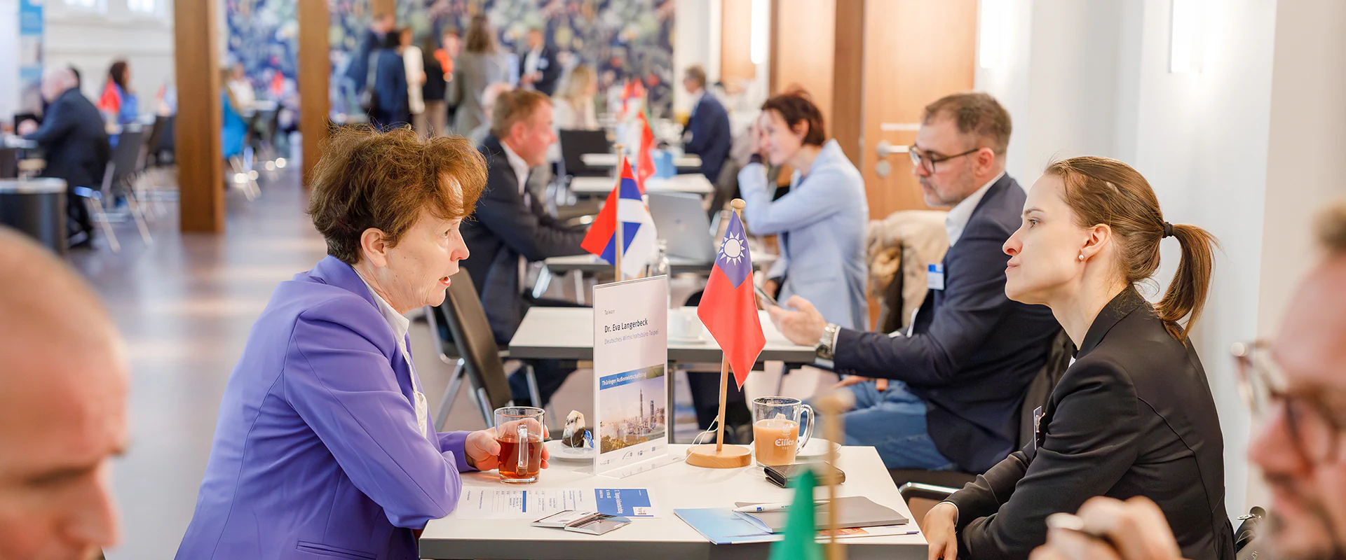 Bildbeschreibung / description: Personen sitzen an Tischen in einer geschäftlichen Meeting‑Umgebung, vor ihnen stehen kleine Länderflaggen und Namensschilder. Im Vordergrund ist ein Tisch mit Dokumenten und einer Flagge zu sehen.