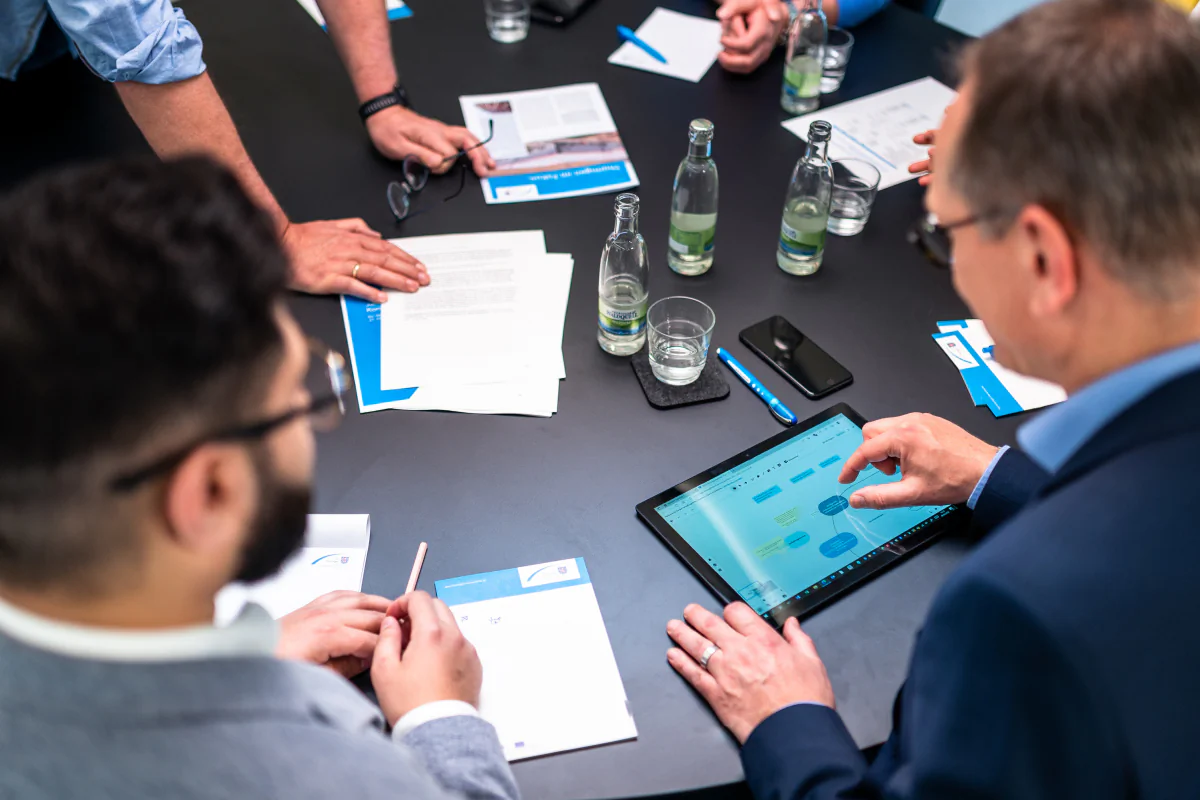 Bildbeschreibung / description: Mehrere Personen sitzen an einem Besprechungstisch und diskutieren Dokumente und ein Tablet mit Diagrammen; auf dem Tisch liegen Wasserflaschen, Gläser, Stifte und Unterlagen.