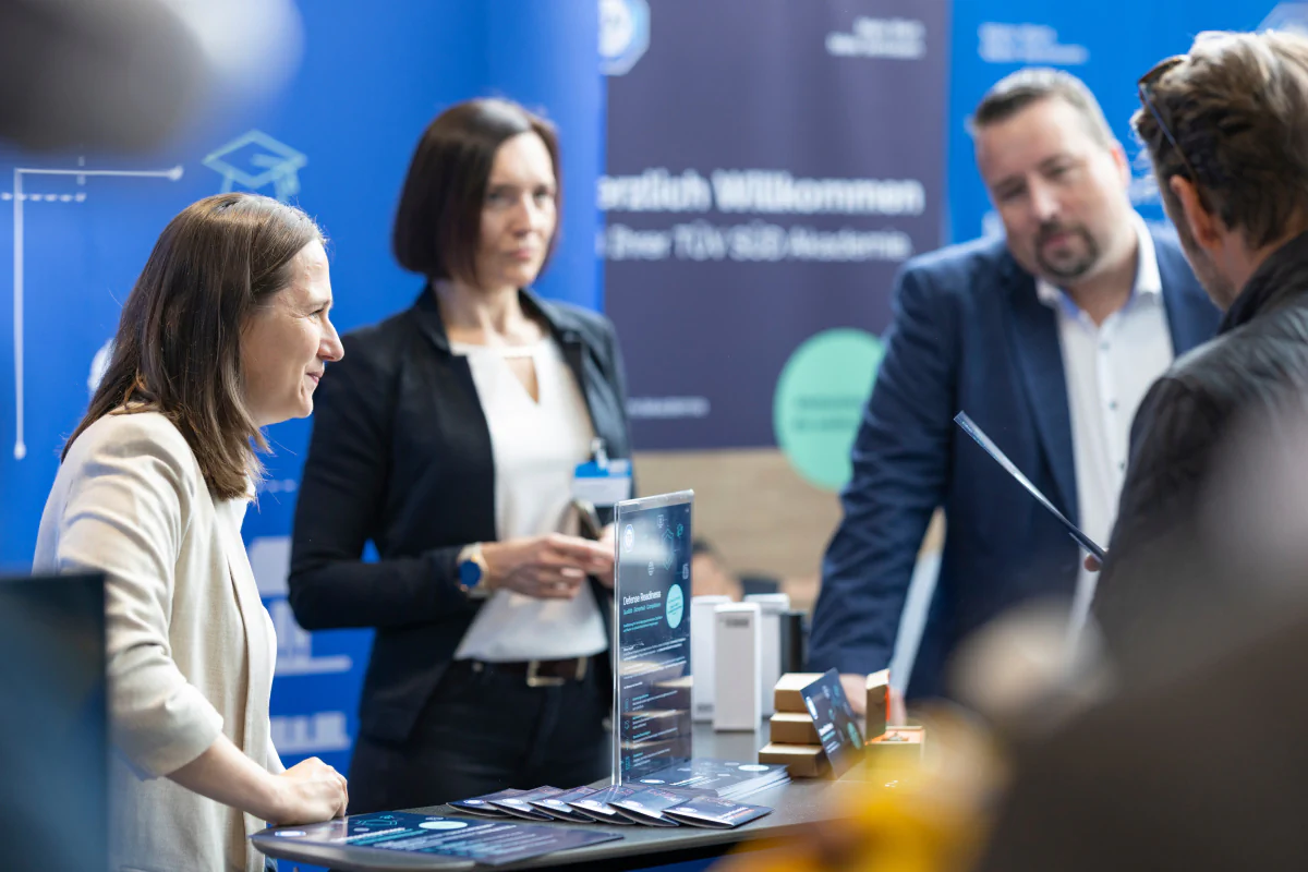 Bildbeschreibung / description: Mehrere Personen stehen an einem Messestand und führen ein Gespräch; im Vordergrund liegen Informationsmaterialien und ein Display, im Hintergrund sind blaue Messegrafiken zu sehen.